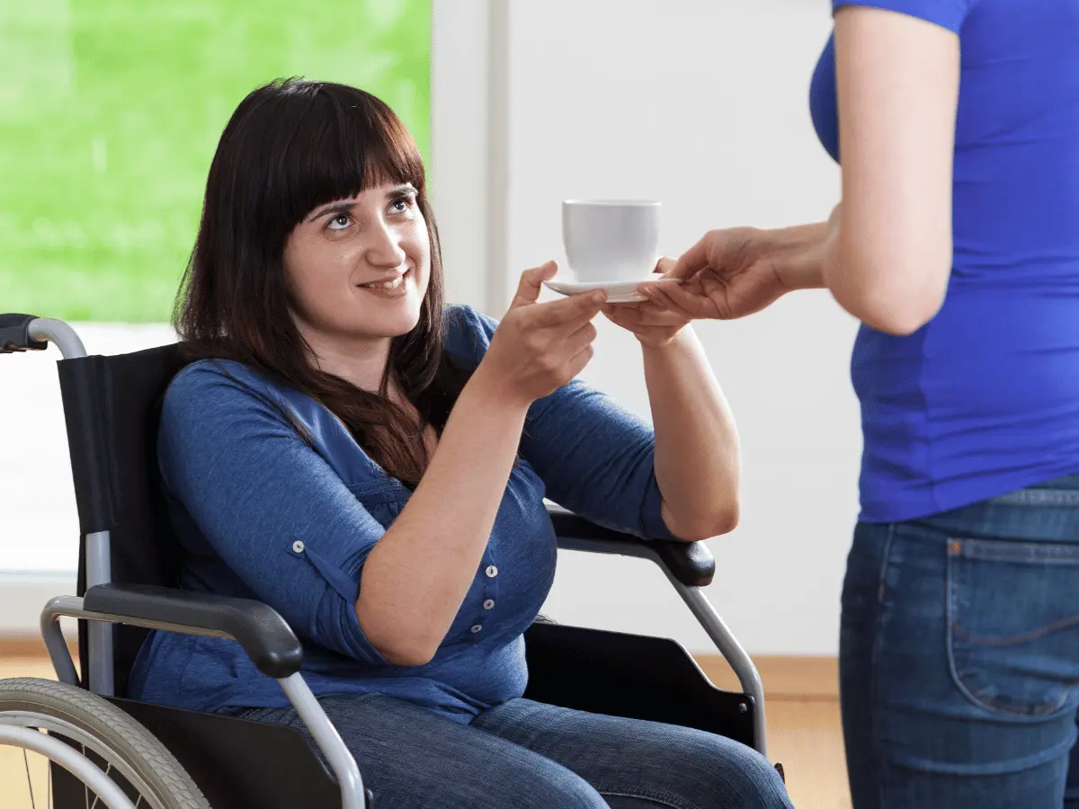Disabled woman in wheelchair taking cup of tea from carer and smiling